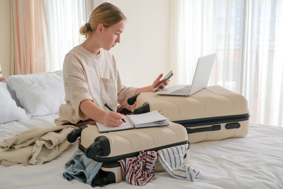 Woman sitting on hotel bed with luggage, taking notes, looking at phone and laptop open